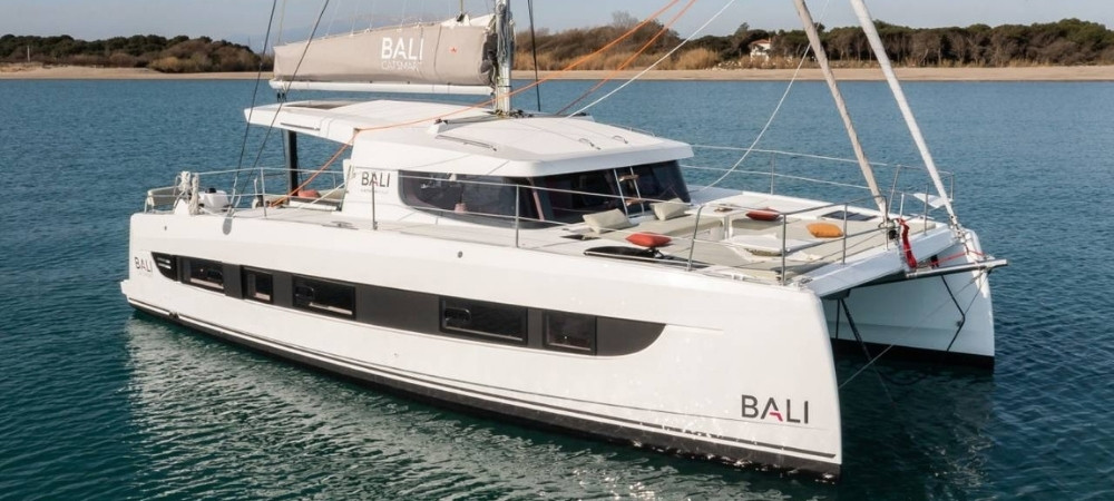 White Bali-brand catamaran on calm blue water near a sandy coastline, with the cabin and deck seating visible.