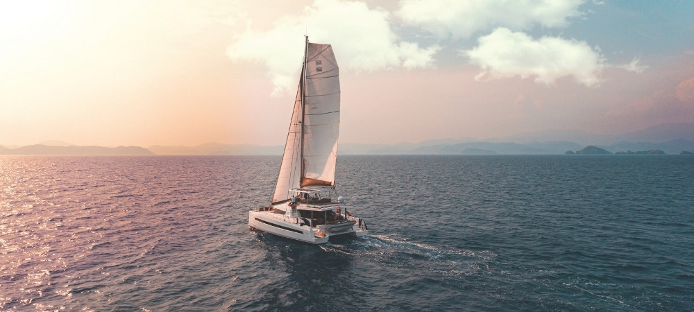 Sailboat gliding across calm ocean at sunset with a pink-orange sky and distant horizon.
