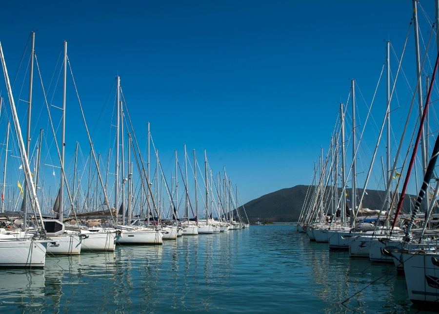 Row of white sailboats docked along a calm harbor with a blue sky and distant hills in the background/side view of a marina with tall masts on both sides.